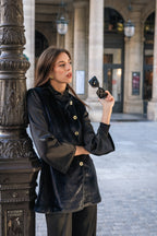 Woman in a black outfit holding sunglasses, standing next to a decorative street light in an urban setting in Paris. Office outfit. Work outfit. business outfit. Women suit.  