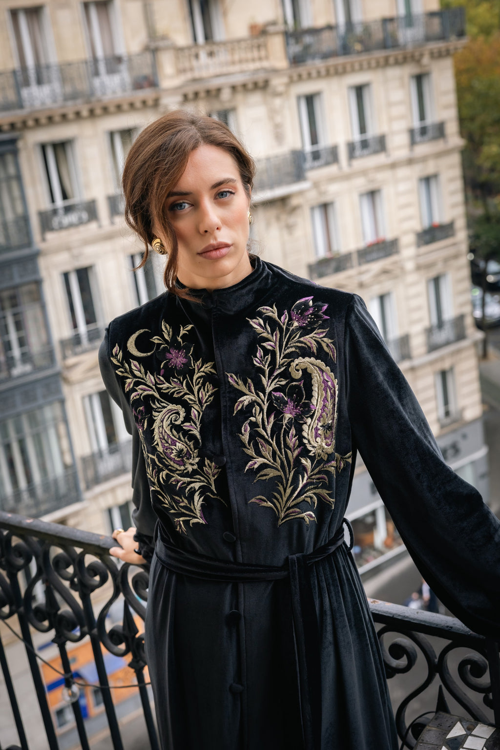 Woman in a black abaya kimono with floral handmade embroidery standing on a balcony with a Parisian cityscape background.