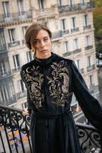 Woman in a black abaya kimono with floral handmade embroidery standing on a balcony with a Parisian cityscape background.