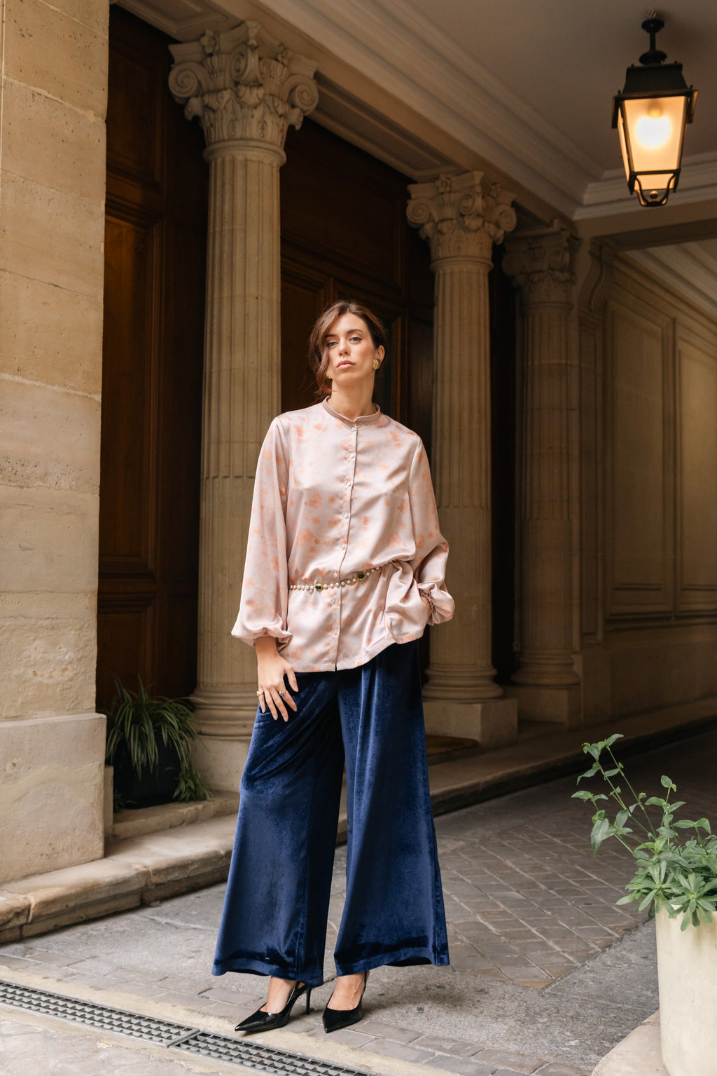 Woman in a pink blouse and blue pants standing in an elegant outdoor setting with columns in Paris.