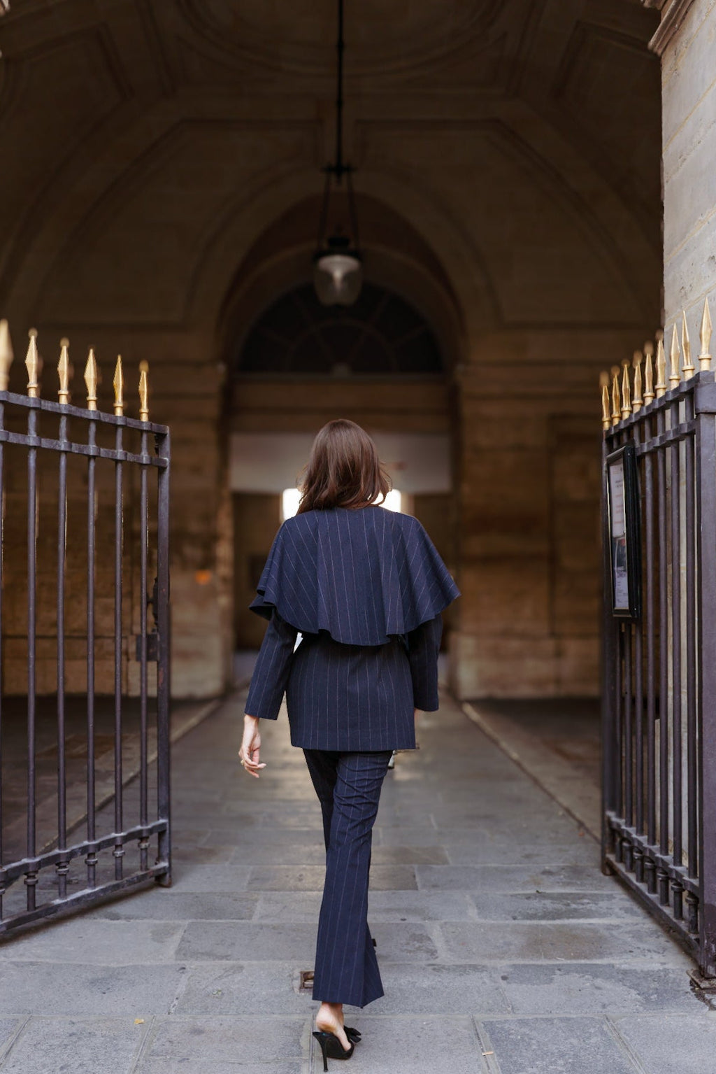 Person in a navy pinstripe suit outfit walking through an archway with stone walls and metal gates.