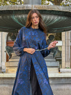Woman in a blue floral dress standing in front of a fountain playing with her pearl belt.